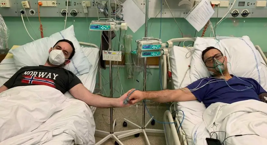 Dwóch mężczyzn w szpitalnych łóżkach, po przeszczepie nerki, trzyma się za ręce. Jeden ma maskę na twarzy, drugi tlenową.