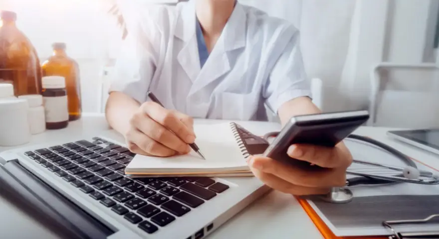 Lekarz w białym kitlu pisze w notatniku, trzymając telefon. Obok laptop, stetoskop i butelki.