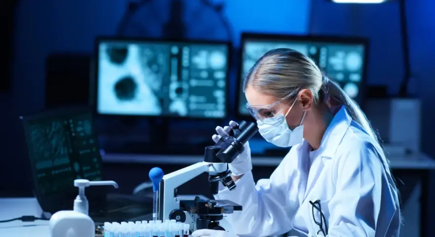Kobieta w masce i okularach ochronnych pracuje przy mikroskopie w laboratorium.