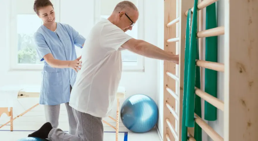 Fizjoterapeutka pomaga pacjentowi ćwiczyć na piłce rehabilitacyjnej przy drabinkach.