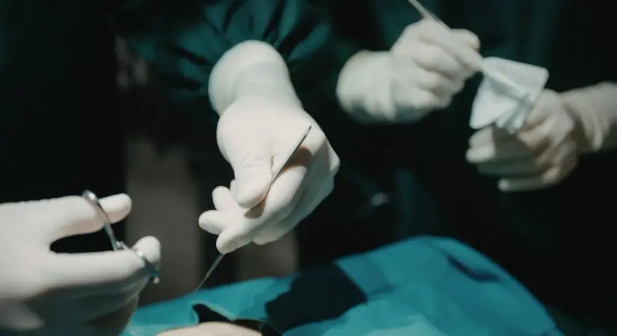 Dłonie chirurga w rękawiczkach trzymające narzędzia chirurgiczne i gazę podczas operacji.