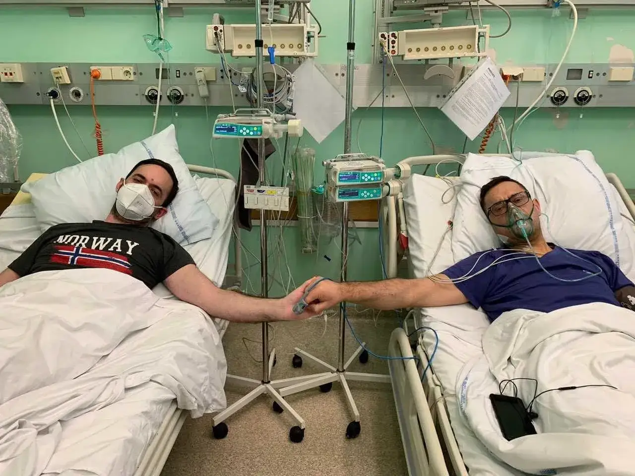 Dwóch mężczyzn w szpitalnych łóżkach, po przeszczepie nerki, trzyma się za ręce. Jeden ma maskę na twarzy, drugi tlenową.