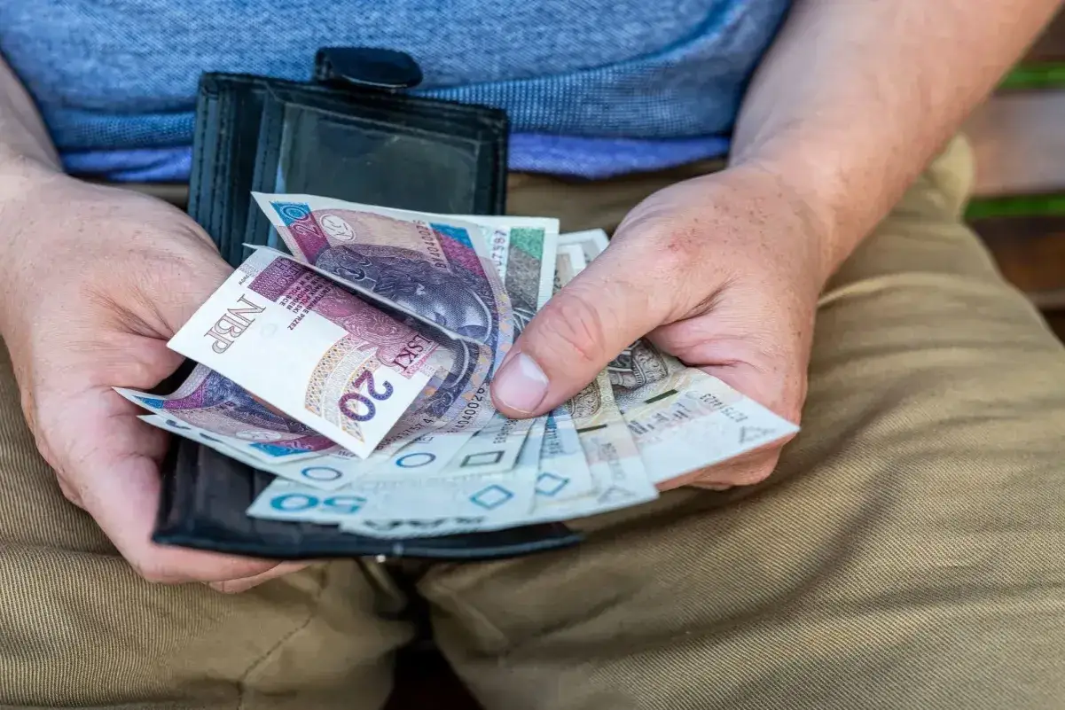 Dłoń trzyma portfel z polskimi banknotami. Może to być dodatek opiekuńczy po 80 roku życia, zapewniający środki na codzienne potrzeby.