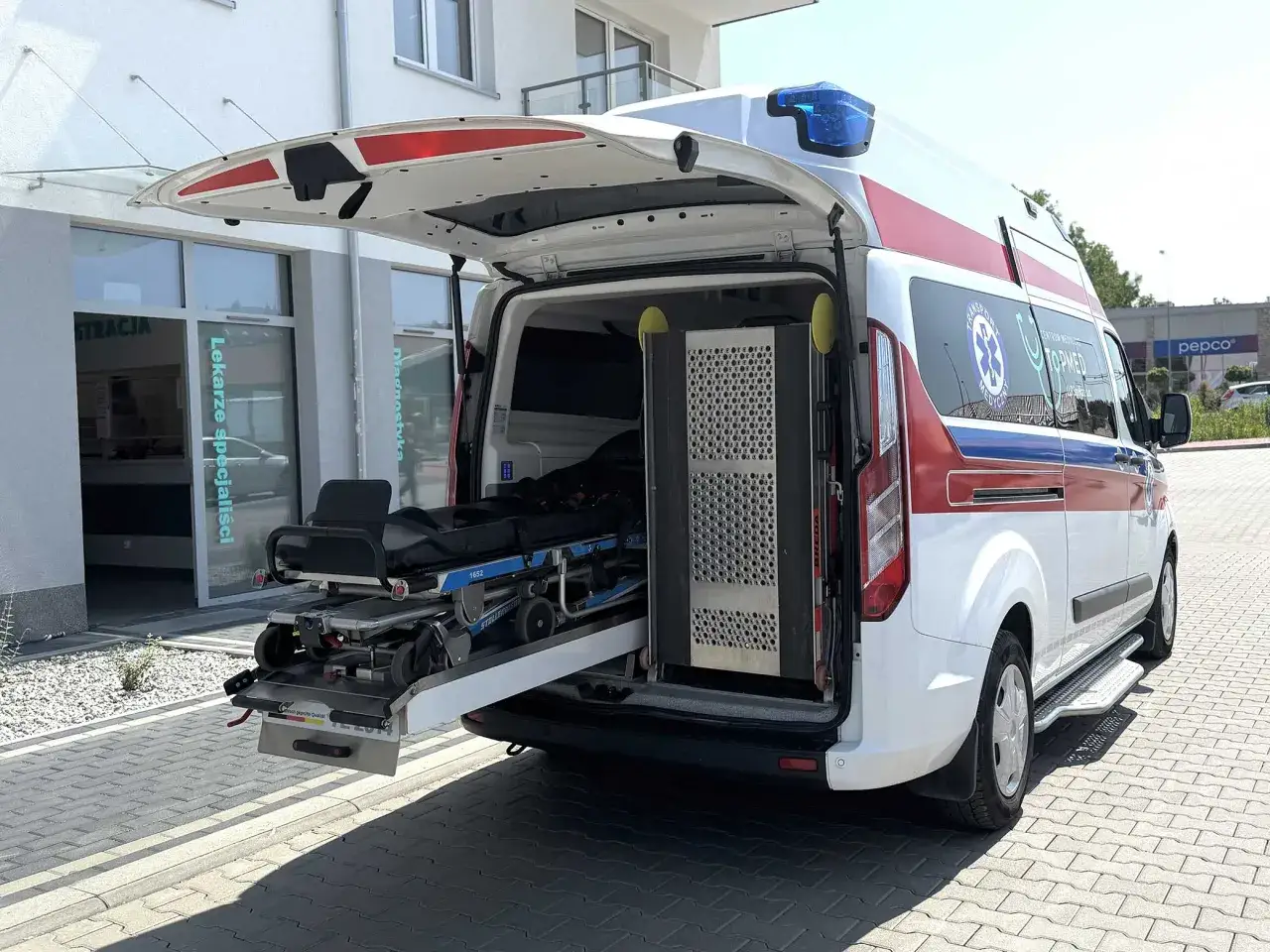 Ambulans z wysuniętymi noszami przed budynkiem z napisem