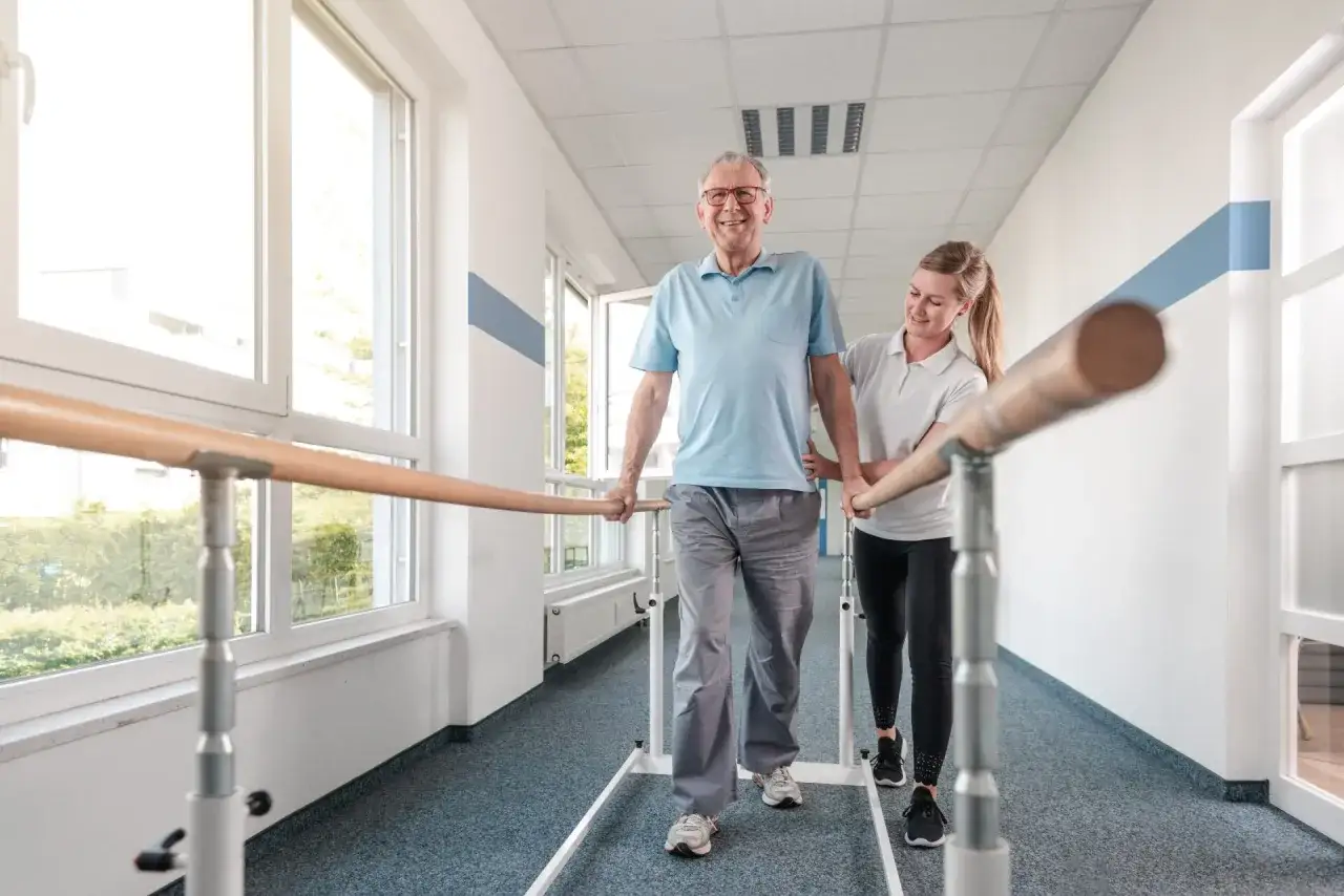 Starszy pan z uśmiechem ćwiczy chód przy poręczach, wspierany przez fizjoterapeutkę. To może być rehabilitacja po skierowaniu, gdzie ZUS wysyła do sanatorium na kręgosłup.